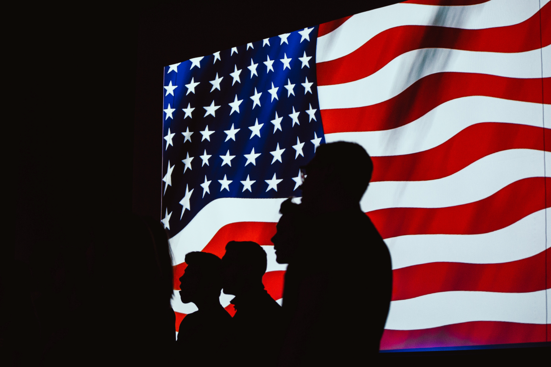 The photo shows the silhouette of four persons standing against the background of the US flag.